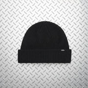 A black ribbed knit beanie hat with a folded cuff, shown against a solid light beige background. A small white Vans logo is visible on the right side of the cuff.